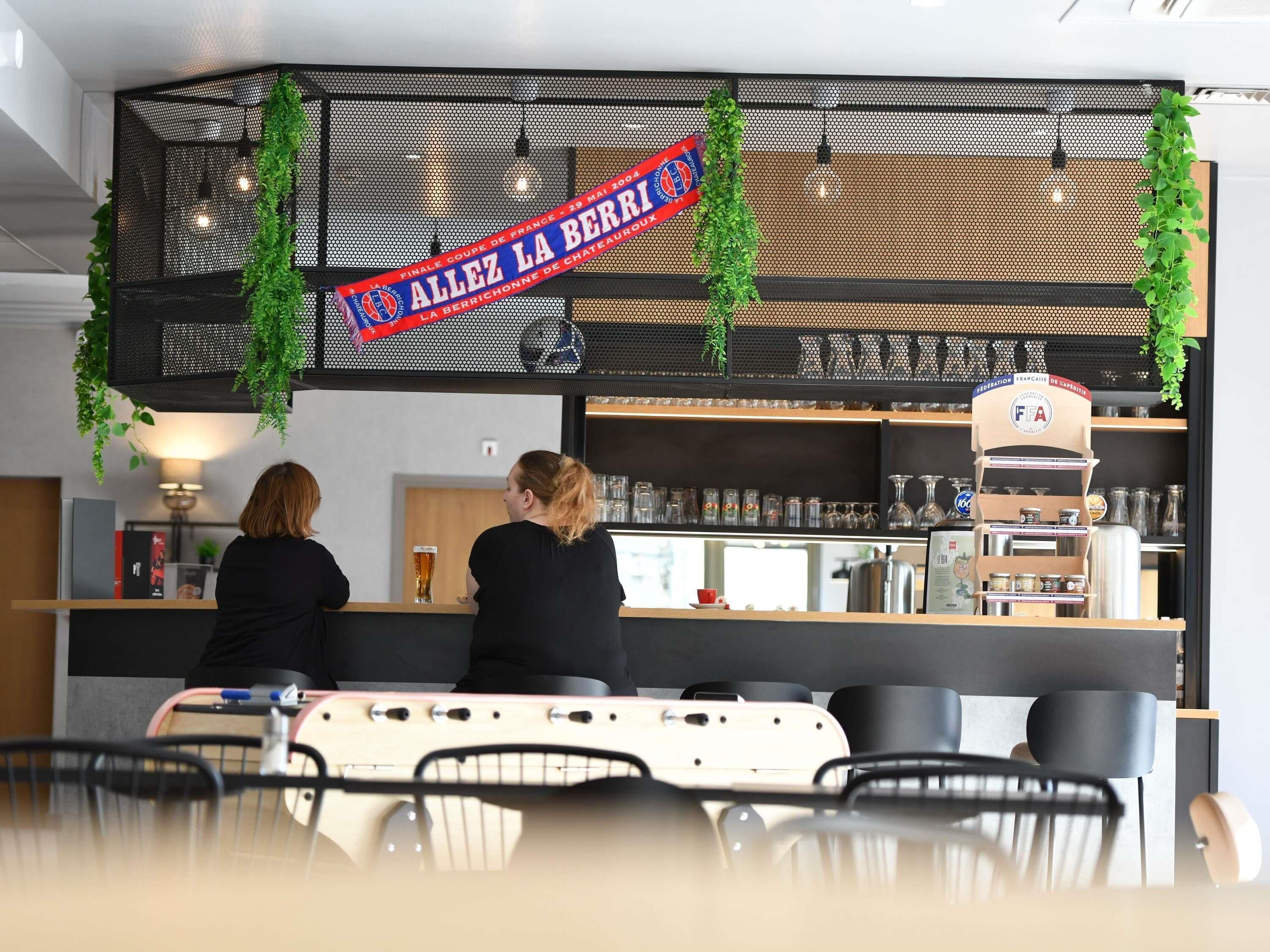 En bardisk med en skylt som visar texten ”ALLEZ LA BERRI”, en disk med glas och en sittgrupp med stolar.