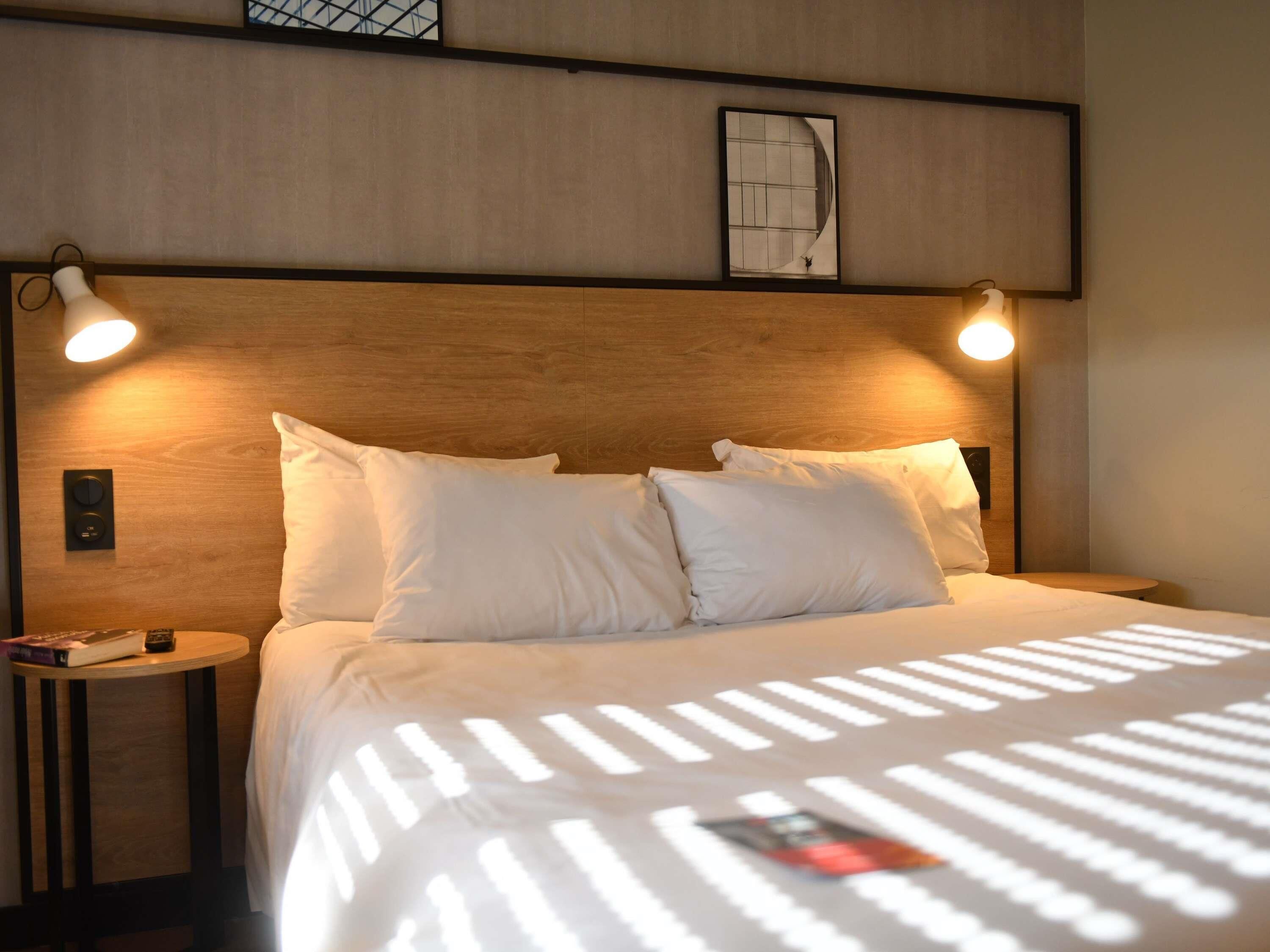 Une chambre d’hôtel avec une tête de lit en bois, deux lampes de chevet, un lit avec du linge de lit blanc et une petite table sur laquelle se trouvent un livre et une télécommande.
