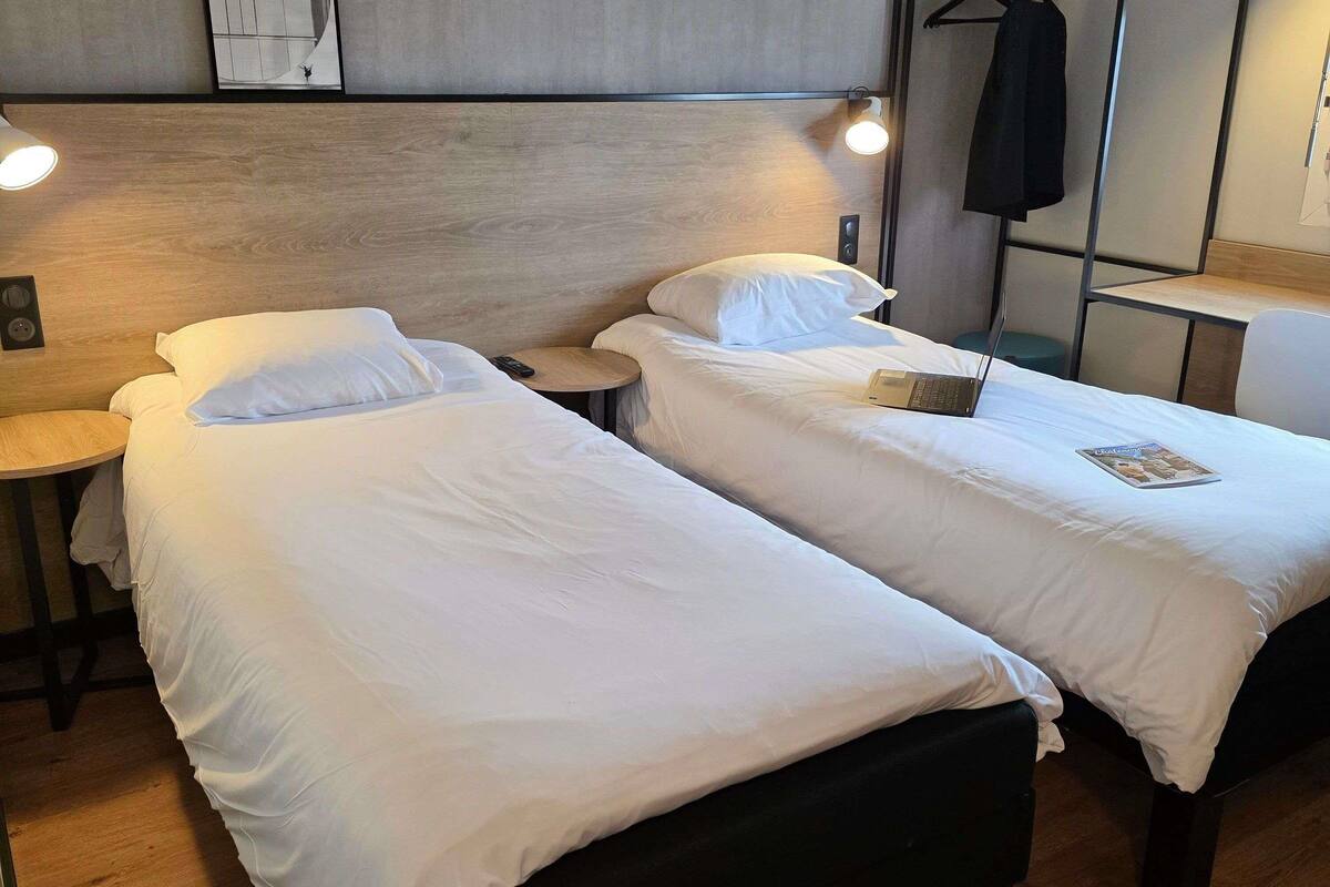 A hotel room with two beds, a small round table, a wall-mounted light, and a coat rack.