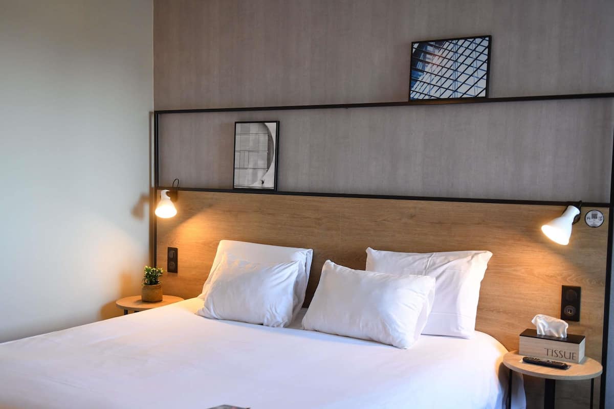A neatly made bed with white linens, two bedside lamps, a small bedside table with a tissue box and remote, and a framed picture on the wall.