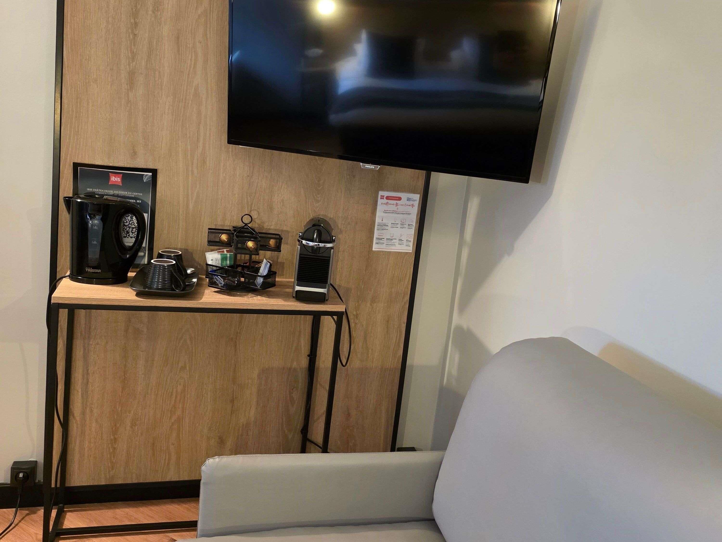 A room with a mounted flat-screen TV, a coffee station with a THESIA coffee maker, a black electric kettle, and a black coffee machine on a wooden table.
