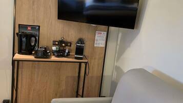 A room with a mounted flat-screen TV, a coffee station with a THESIA coffee maker, a black electric kettle, and a black coffee machine on a wooden table.