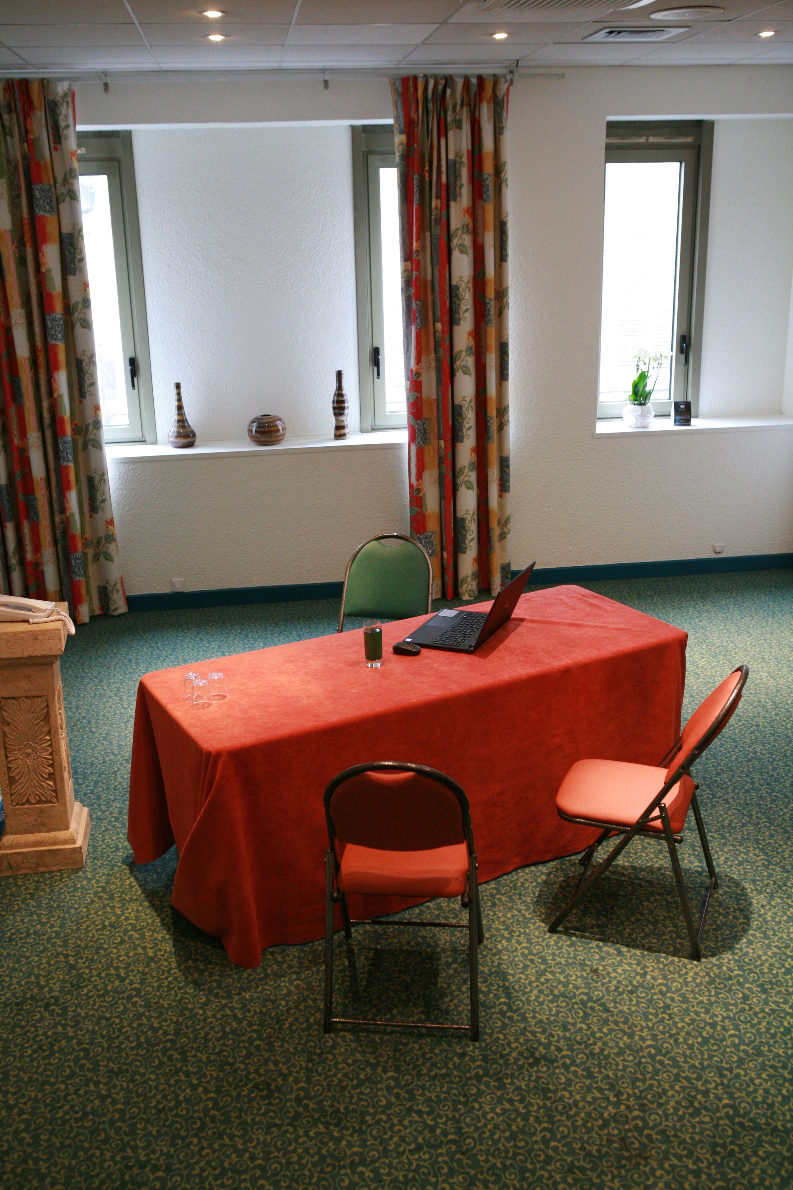 Uma sala de conferências com uma mesa vermelha, duas cadeiras, um laptop e um vaso no parapeito da janela.