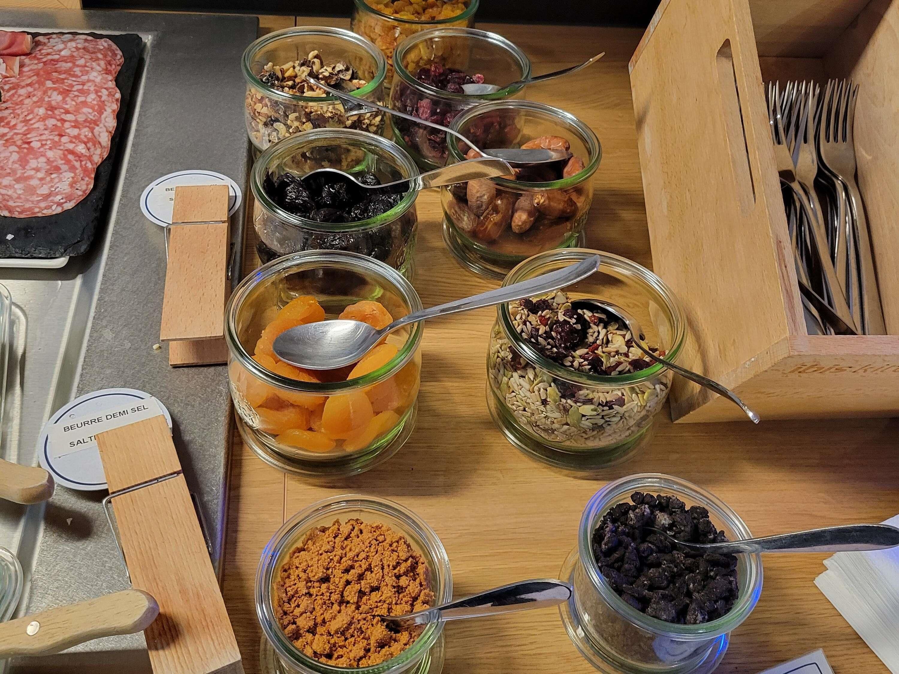 A variety of food items in glass jars on a wooden table, including dried fruits, nuts, and other snacks.