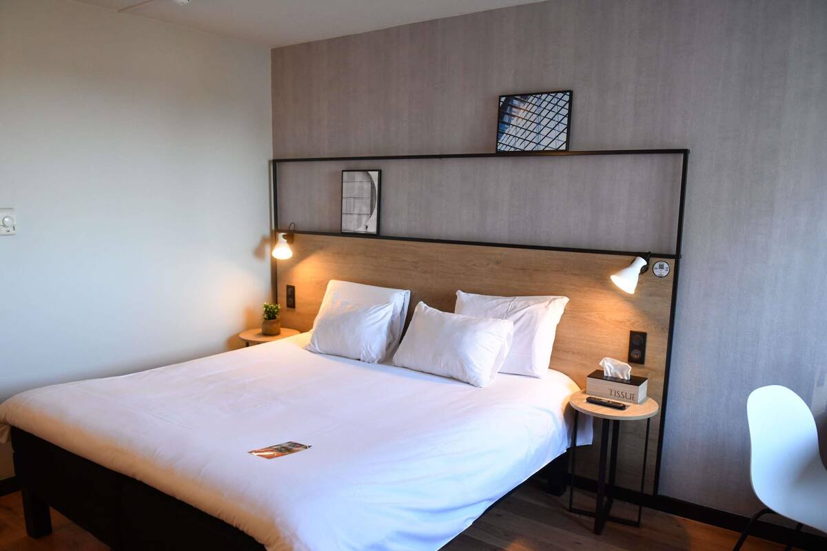 A neatly made bed with white linens, a bedside table with a tissue box, and a modern chair.