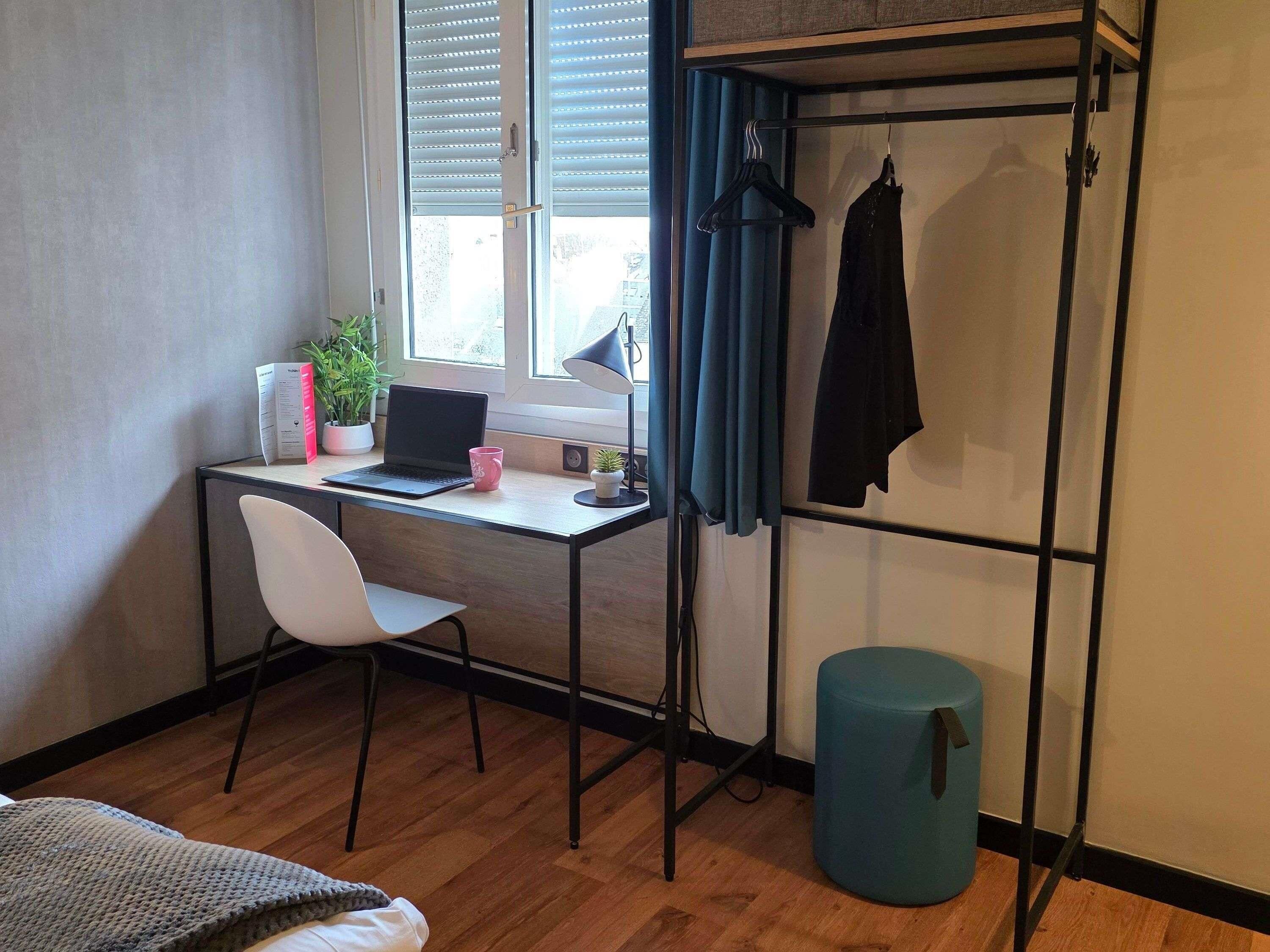 Habitación compacta con escritorio de madera, una computadora portátil, una silla blanca, una pequeña planta en maceta y un perchero.