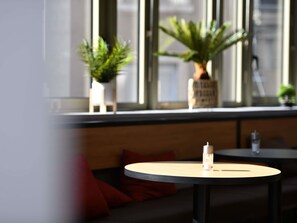 A seating area with a round table and a bench, a small potted plant, and a window with blinds. - ibis Chateauroux (Chateauroux)