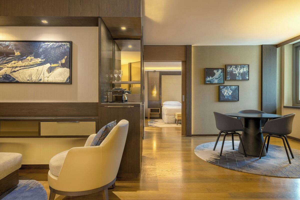 A modern hotel room with a wooden floor, a dining area with a round table and chairs, a sofa, and a large painting on the wall.