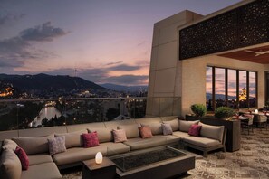 A rooftop terrace with a sofa, a table, and chairs, overlooking a cityscape and a river at dusk. - Sheraton Grand Tbilisi Metechi Palace (Tbilisi)