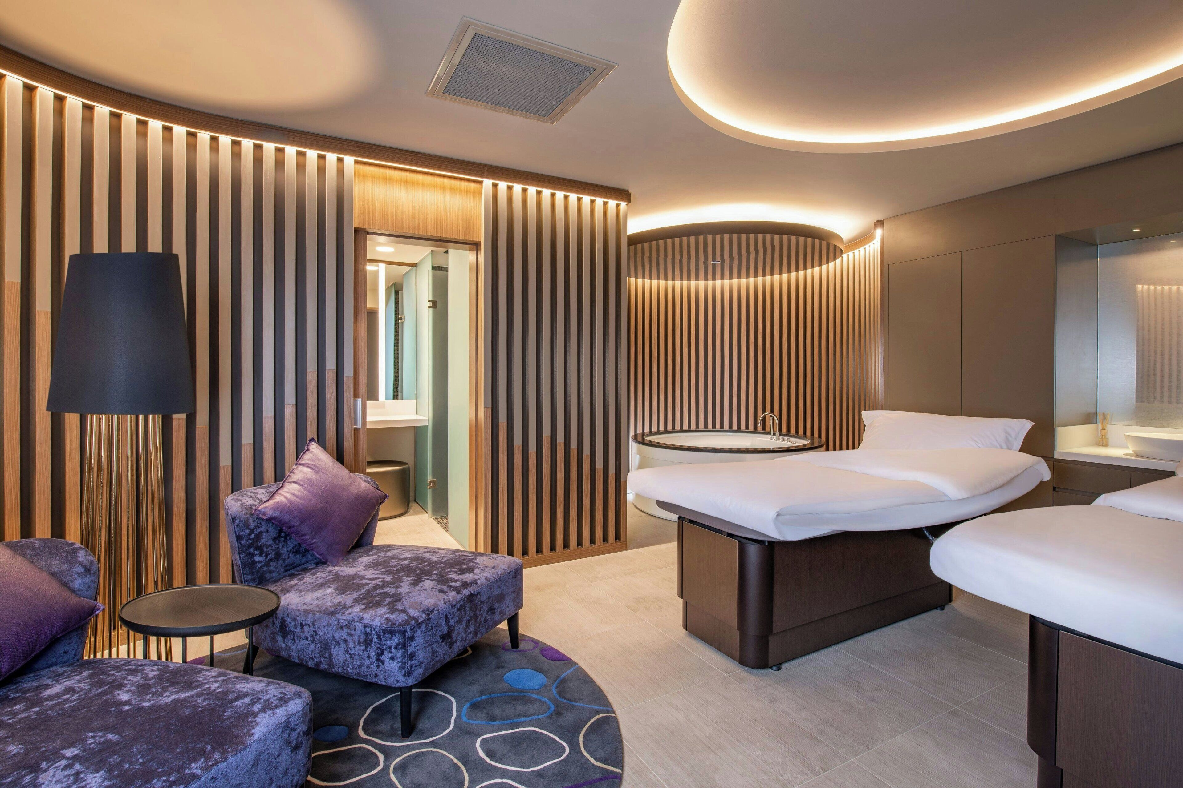 A modern hotel room with a large bed, a sitting area with a sofa and ottoman, a small round table, and a large mirror.