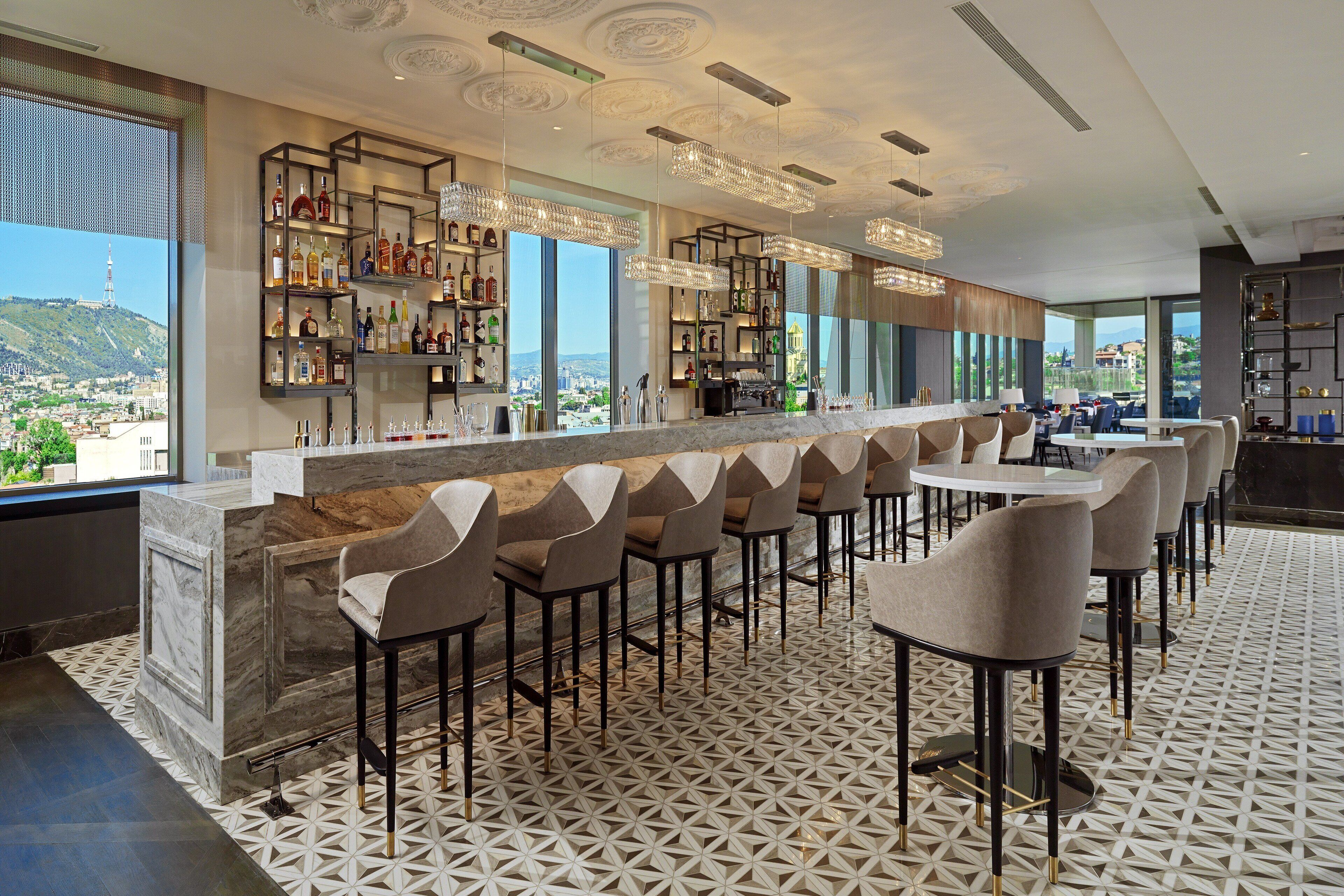 A modern bar area with a marble counter, multiple bar stools, and a view of the cityscape through large windows.