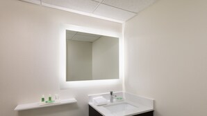 A bathroom with a white sink, a mirror, a toilet, and a shelf with toiletries. - Holiday Inn Clark - Newark Area by IHG (Clark)