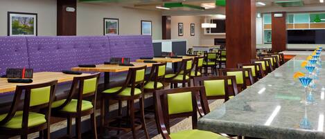 A hotel restaurant with multiple dining tables, green cushioned chairs, a long bar with blue drinks, and a purple booth area.