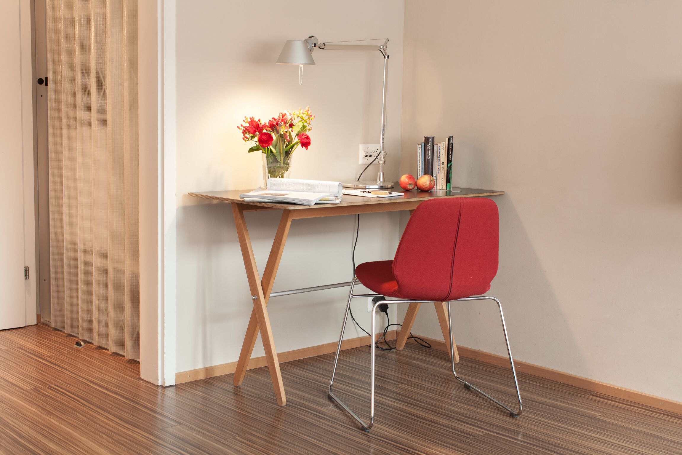 Un espacio de trabajo moderno con un escritorio de madera, una silla roja, una lámpara de escritorio, un jarrón con flores y algunos libros y manzanas.