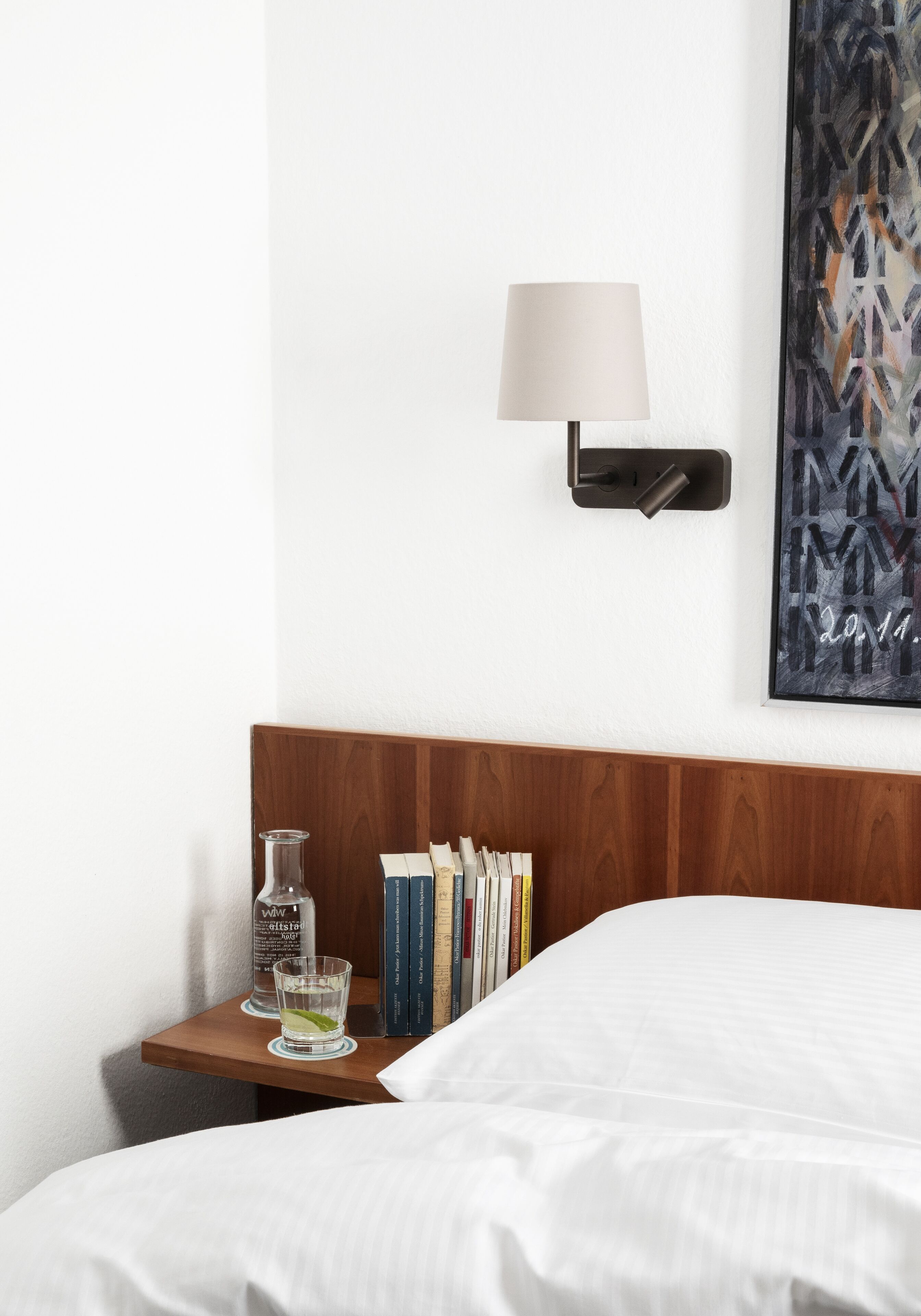 A neatly made bed with white linens, a wooden nightstand with a lamp, a glass of water, and a bottle of water.