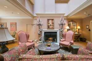 Cozy up by the fireplace in this elegantly furnished living room with a high ceiling, featuring a large coffee table, armchairs, and a sofa.