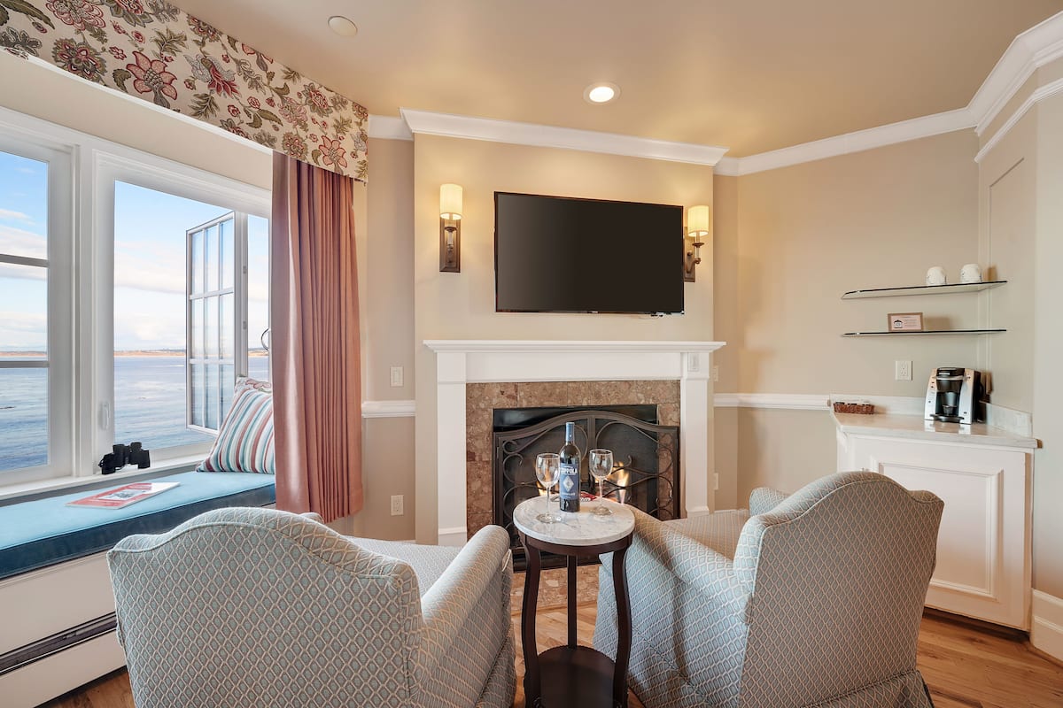 Una sala de estar acogedora con chimenea, un televisor montado encima y un rincón de descanso con dos sillones estampados y una mesita.