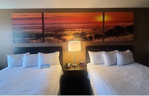 Two single beds with white linens and pillows, a bedside table with a lamp and clock, and a large panoramic painting above.