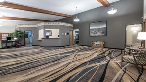 Un hall d’entrée spacieux, doté d’un tapis à motifs, de poutres en bois, d’un comptoir de réception et d’un grand tableau encadré au mur.