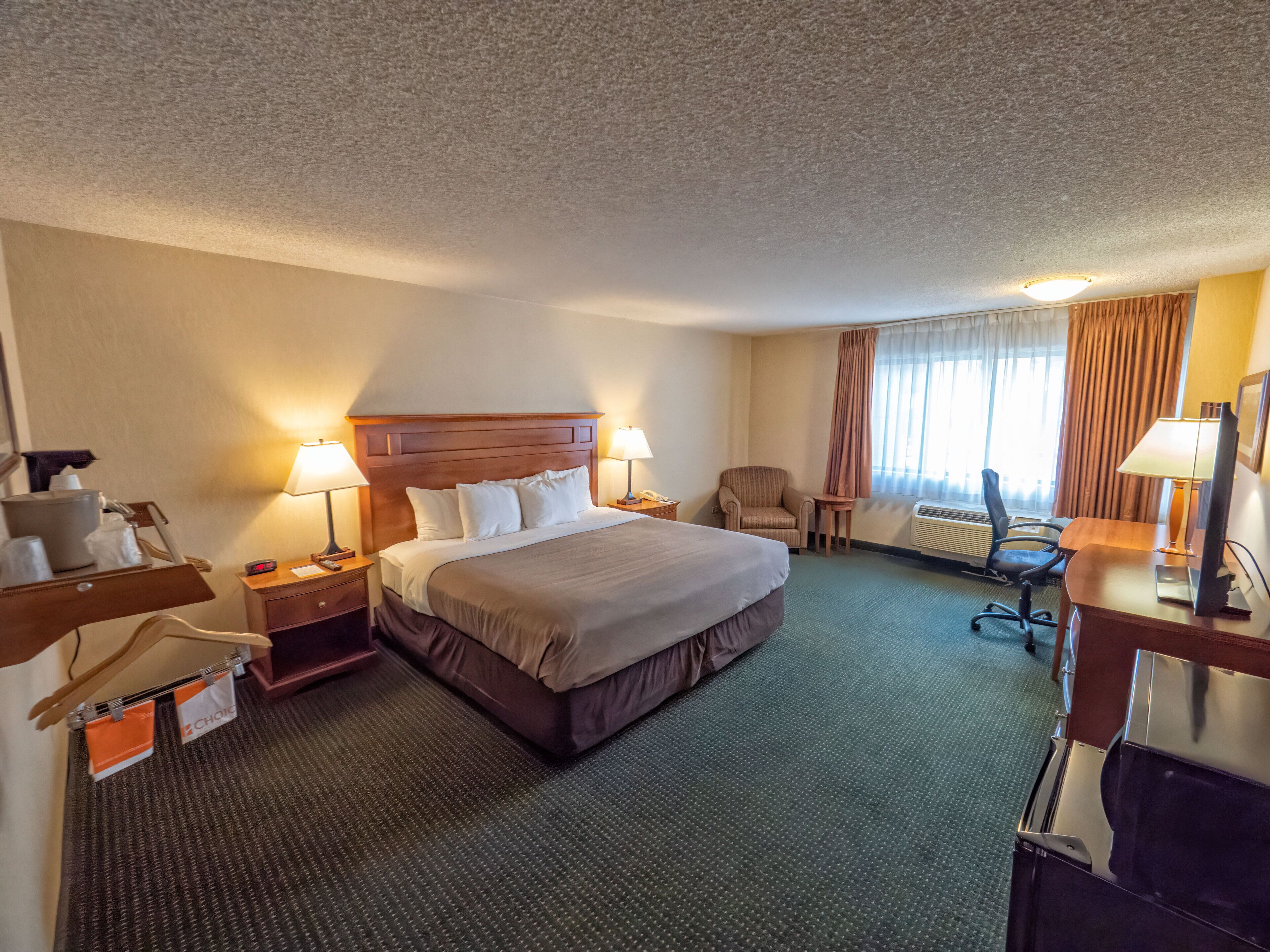 A hotel room with a large bed, two bedside tables with lamps, a chair, a desk with a computer, and a window with curtains.