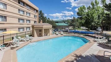 A rectangular pool with lounge chairs and umbrellas, surrounded by a fence and a building with multiple windows.