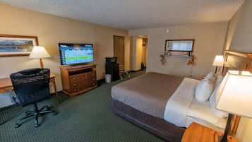 Quarto de hotel com cama, escrivaninha, cadeira, televisão e pequena área de kitchenette.