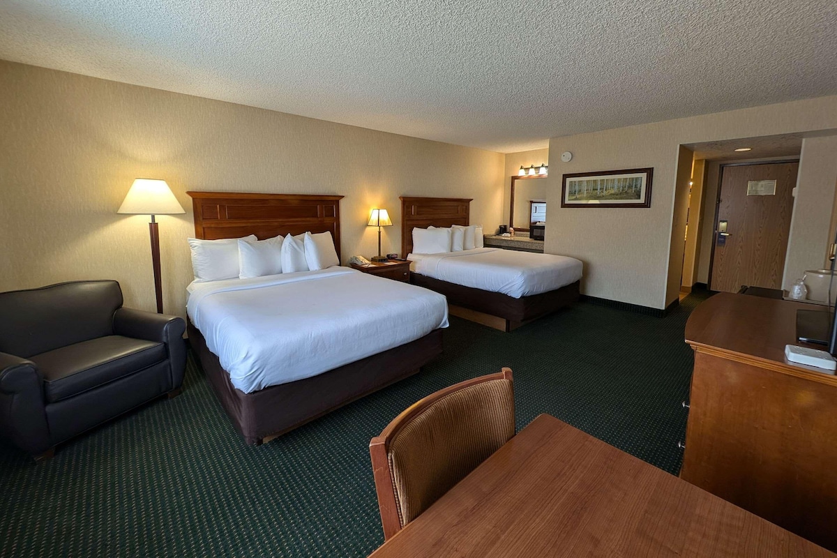 A hotel room with two beds, a desk, a chair, a sofa, and a desk lamp.