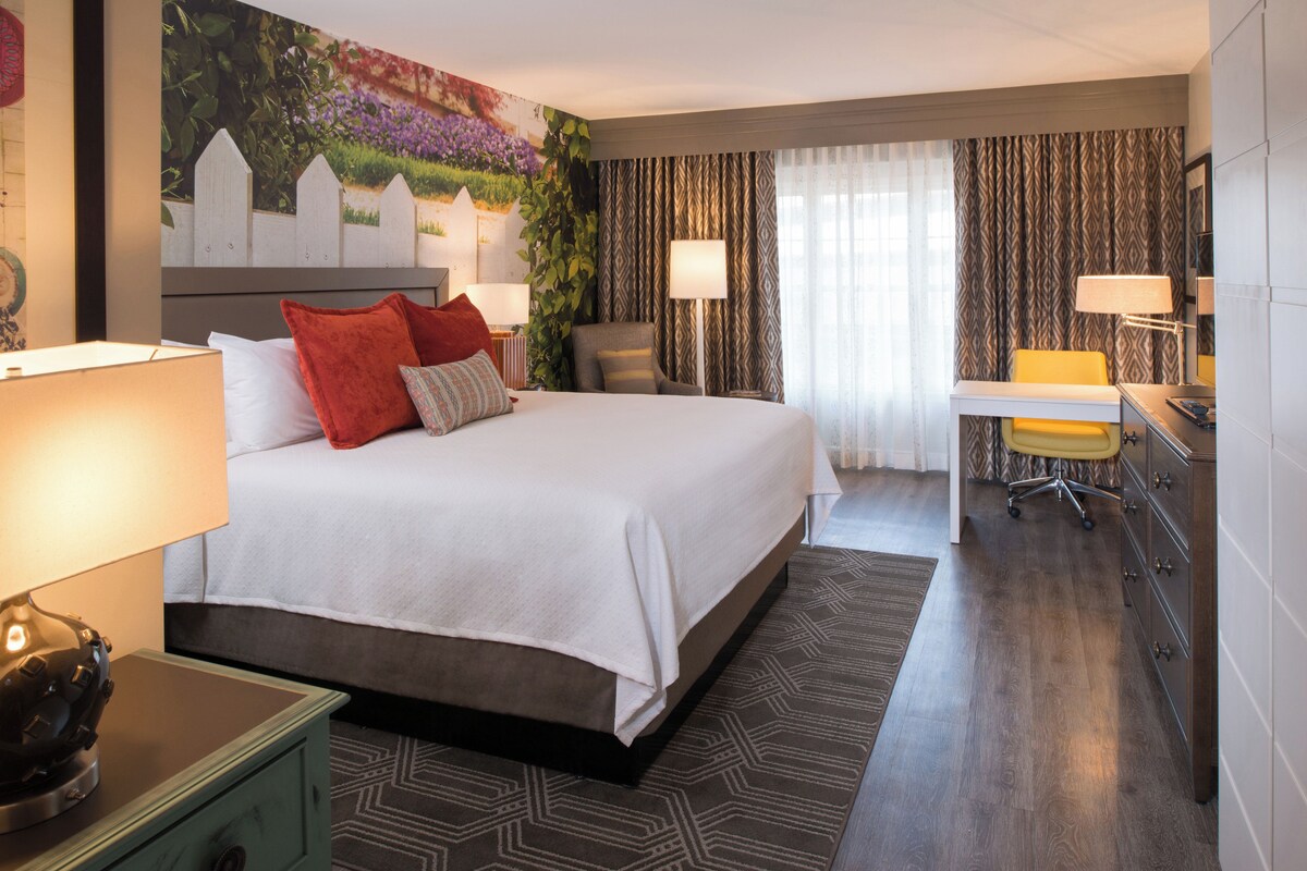 Habitación de hotel con una cama grande, un escritorio, una silla, una lámpara de escritorio, una mesita de noche y una pared con un mural floral.
