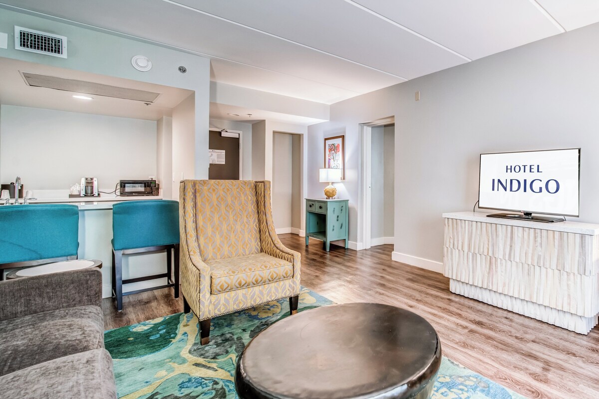 A modern hotel room with a kitchenette, a patterned armchair, a round coffee table, and a television on a white cabinet.
