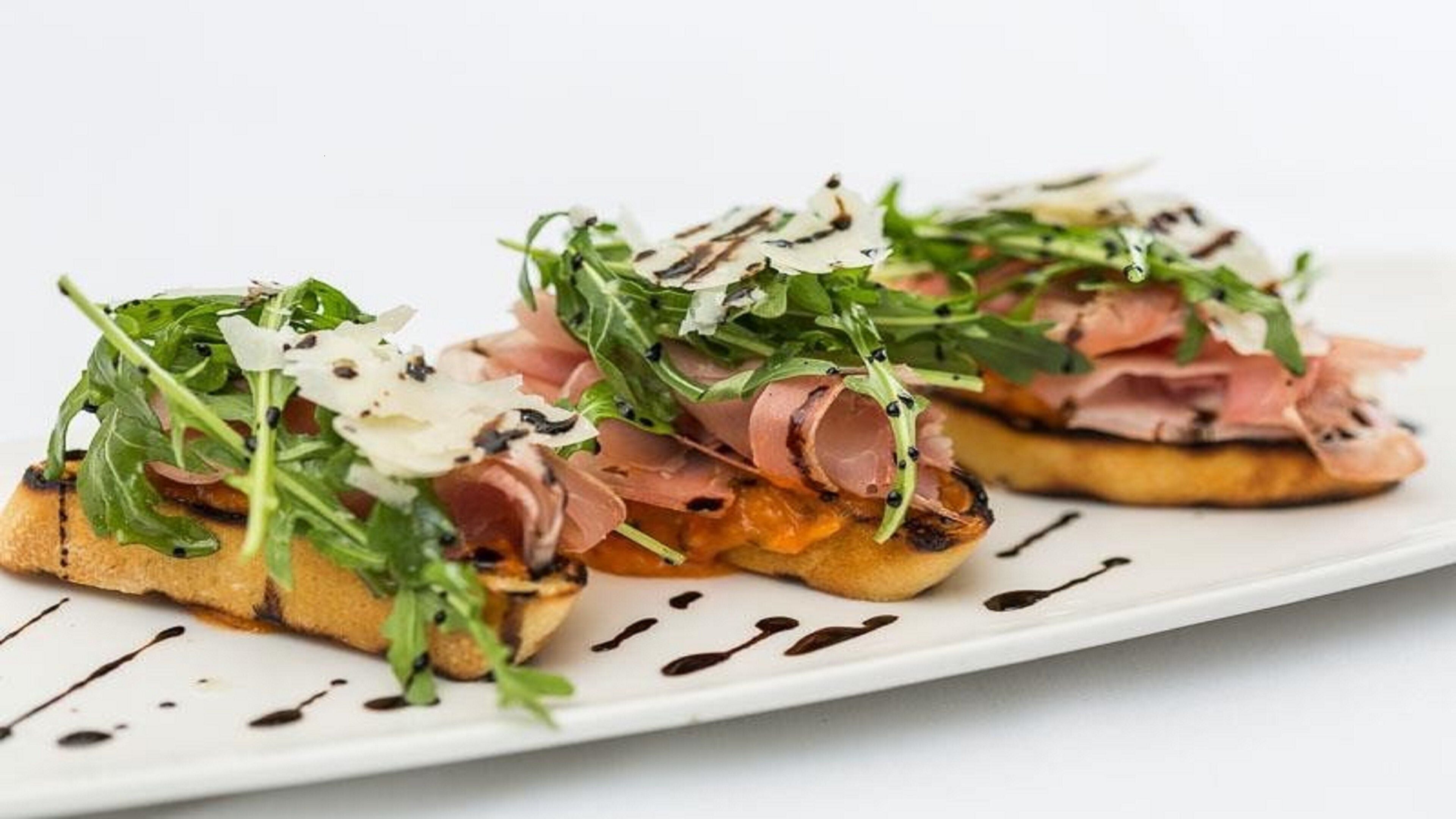 A plate with two pieces of toast topped with greens, prosciutto, and shaved cheese, with a drizzle of sauce.