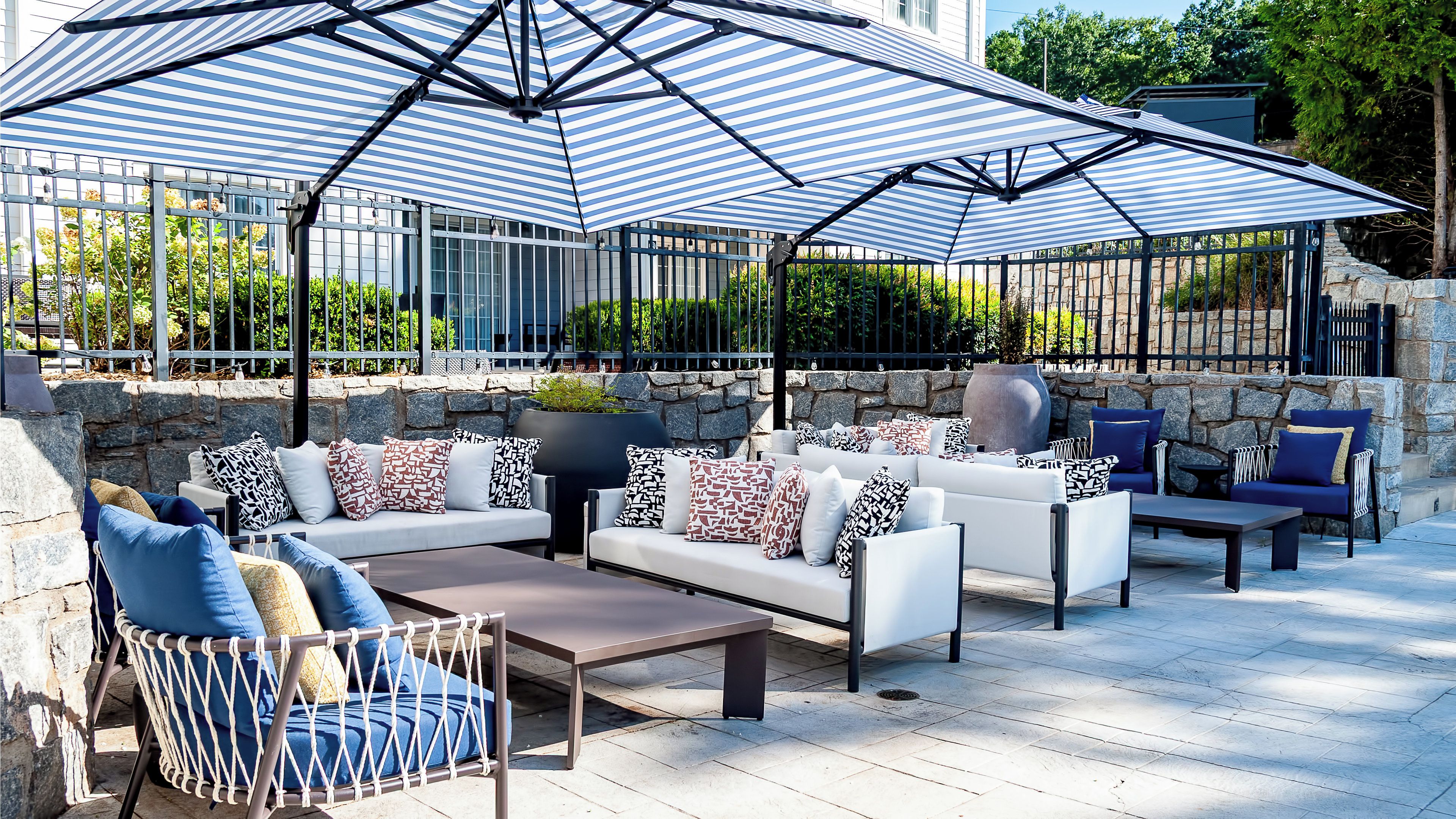 Een terras met een achtergrond van een stenen muur, een grote gestreepte parasol en verschillende zitplaatsen, waaronder banken en stoelen.