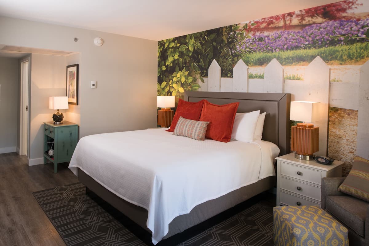 Habitación de hotel ordenada con una cama grande, dos mesitas de noche con lámparas, un pequeño rincón de descanso y un mural en la pared.