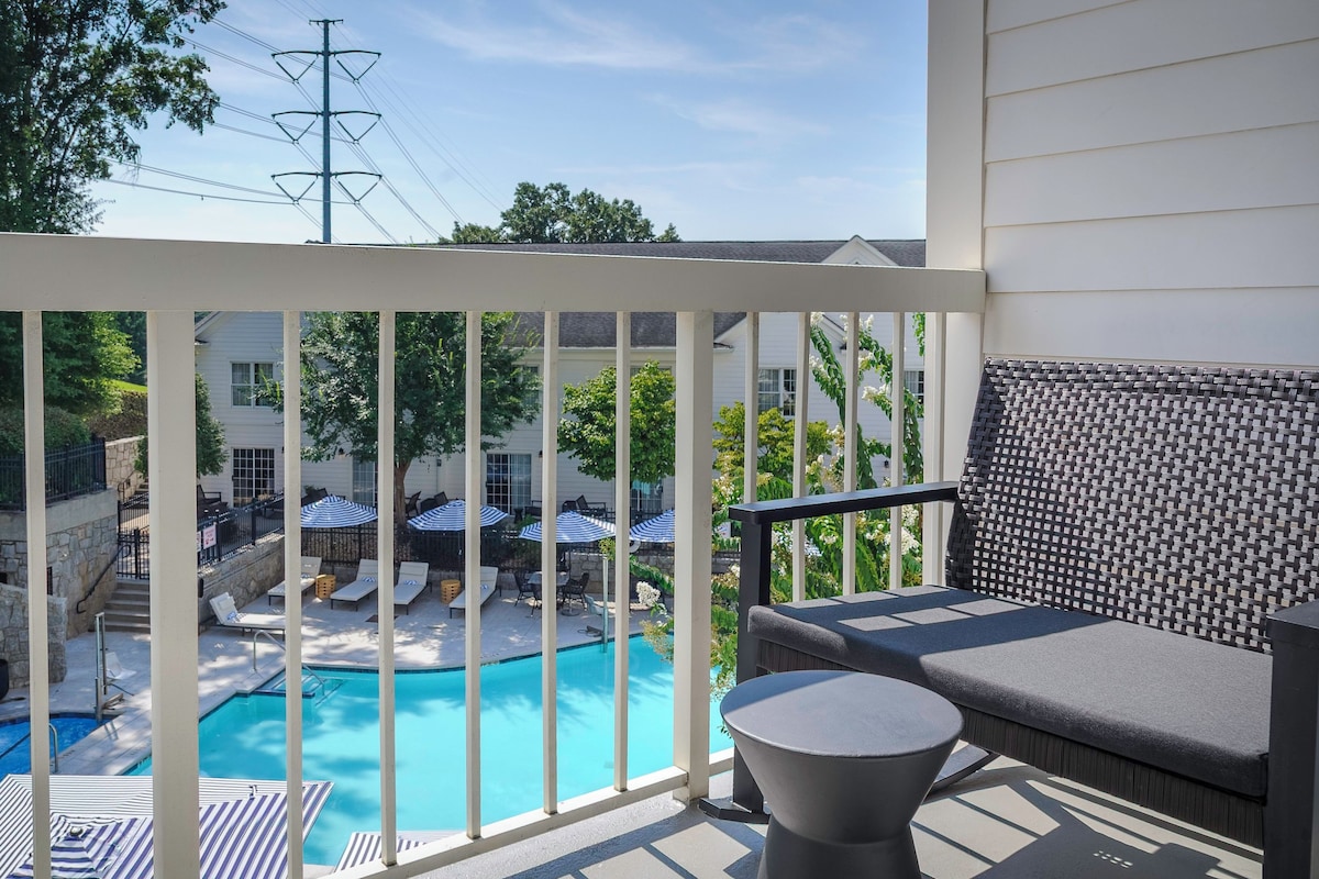 A balcony with a view of a swimming pool, outdoor seating, and a stone waterfall feature.