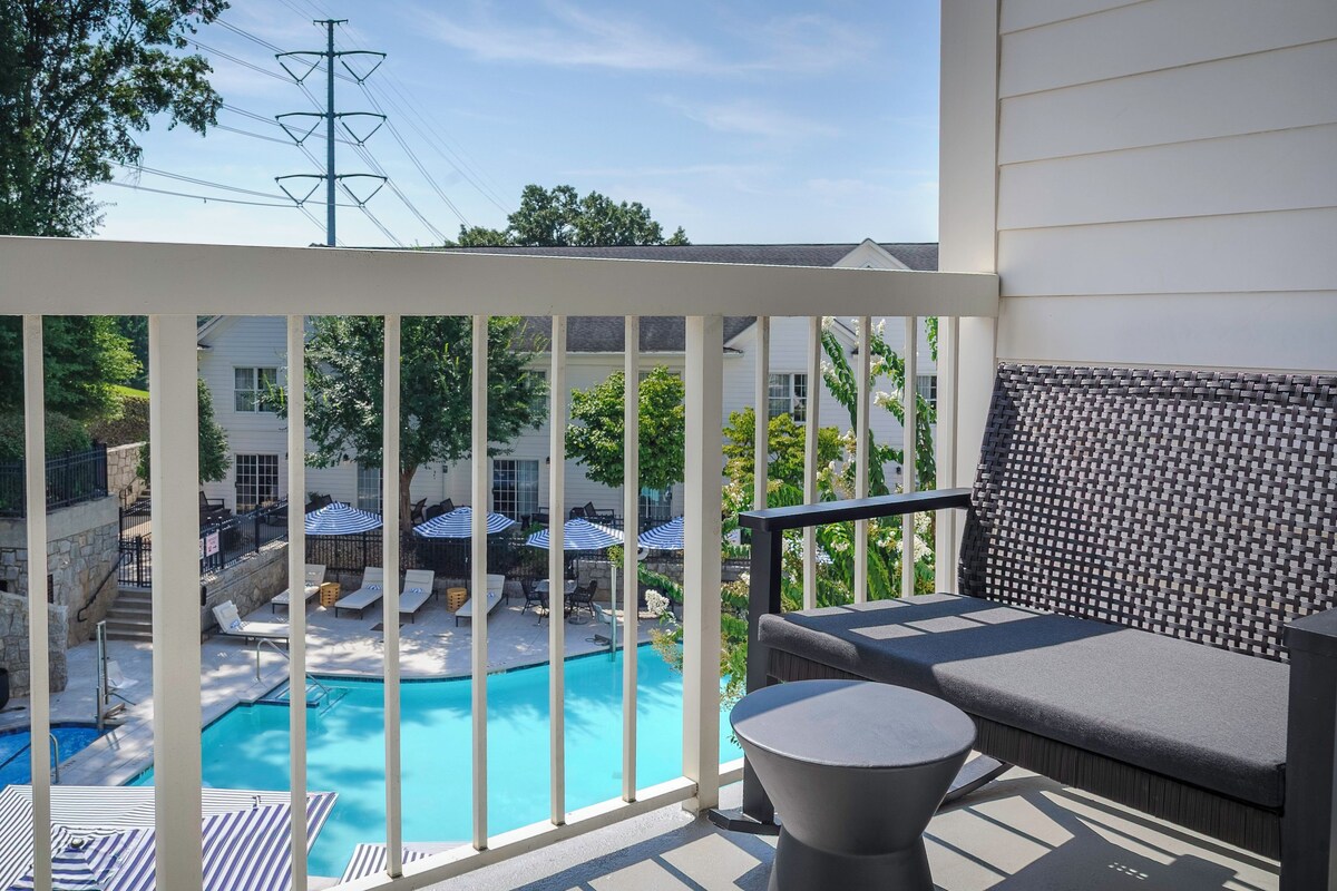 A balcony with a view of a swimming pool, outdoor seating, and a stone waterfall feature.