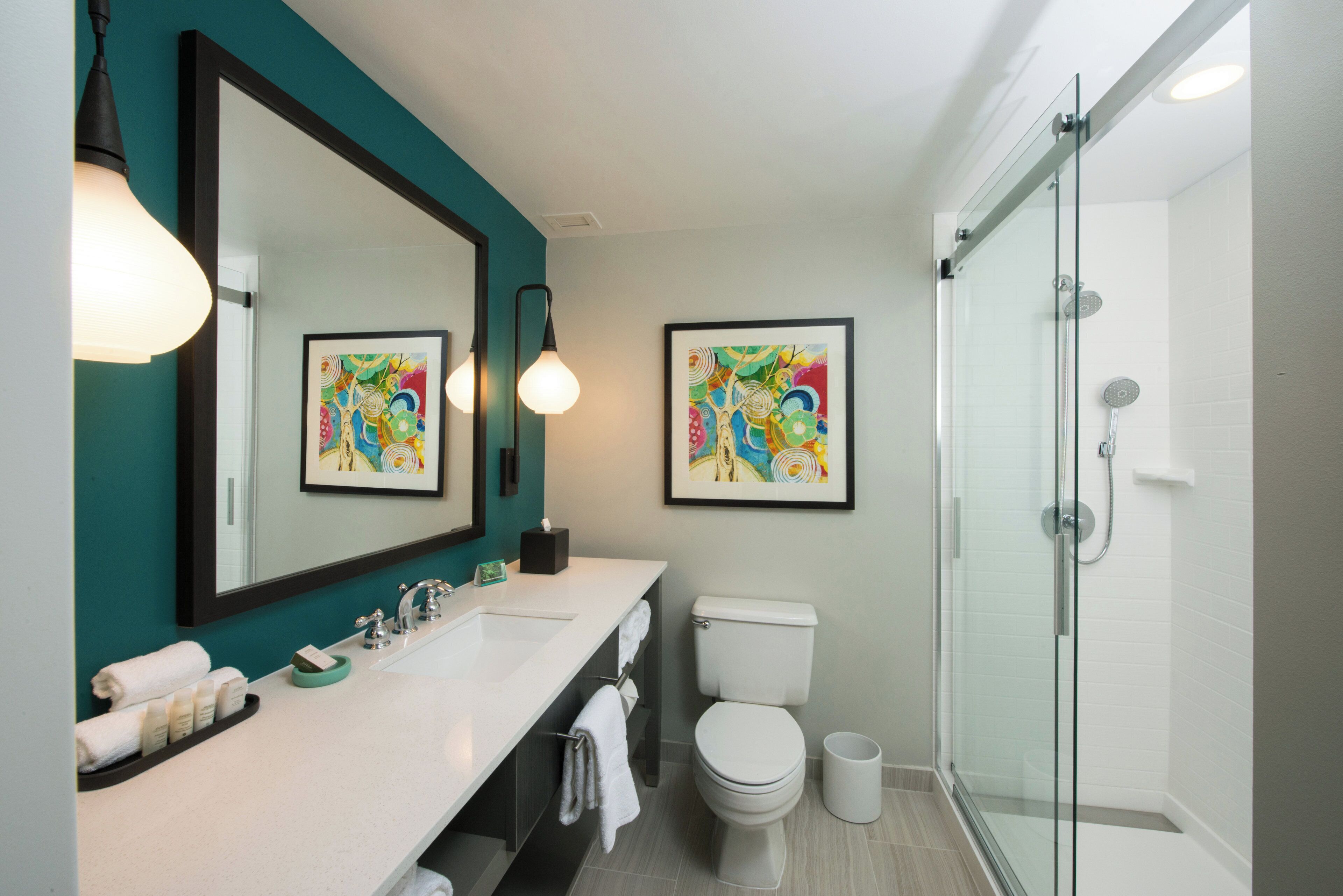 A modern bathroom with a glass shower enclosure, a white sink with a mirror, and two framed abstract paintings on the wall.
