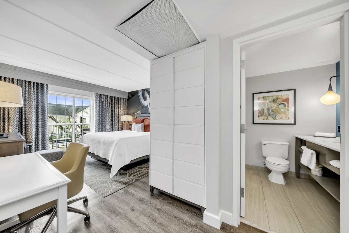 A hotel room with a bed, desk, chair, and a bathroom with a toilet and sink.