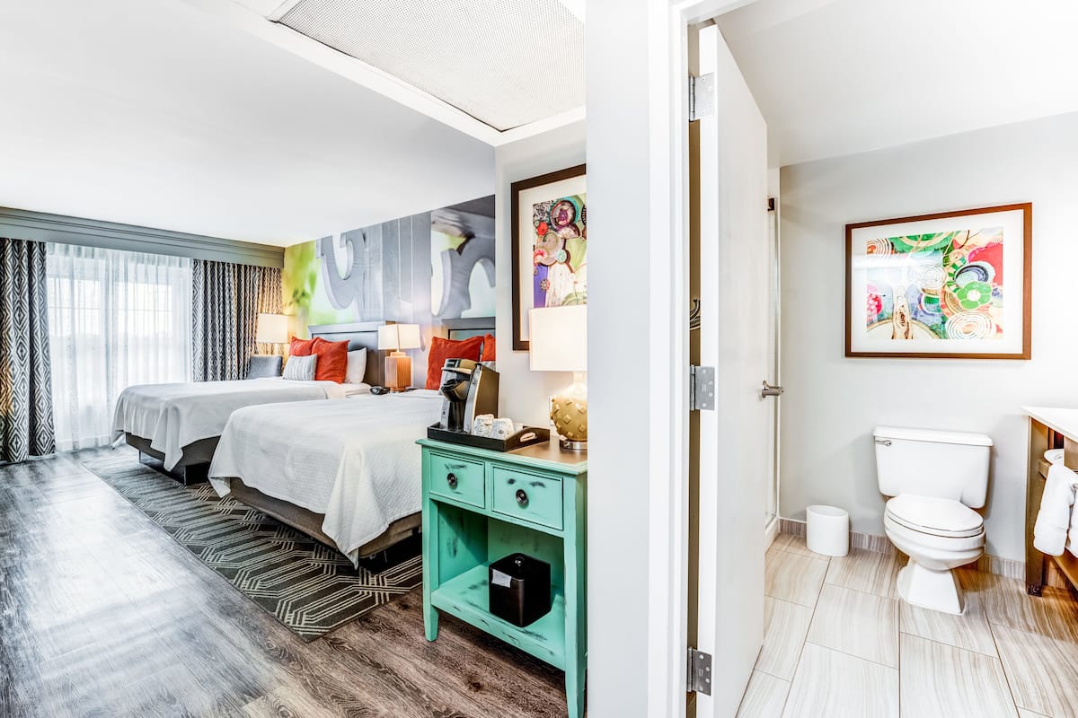 Habitación de hotel con dos camas, una mesita de noche color aguamarina, un baño con inodoro y lavabo, y obras de arte en las paredes.