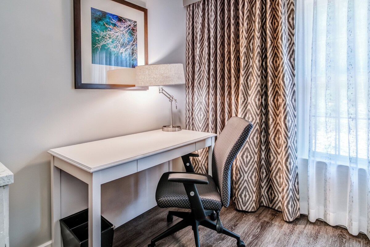 A hotel room with a white desk, a black office chair, a framed artwork on the wall, patterned curtains, and a window with sheer curtains.