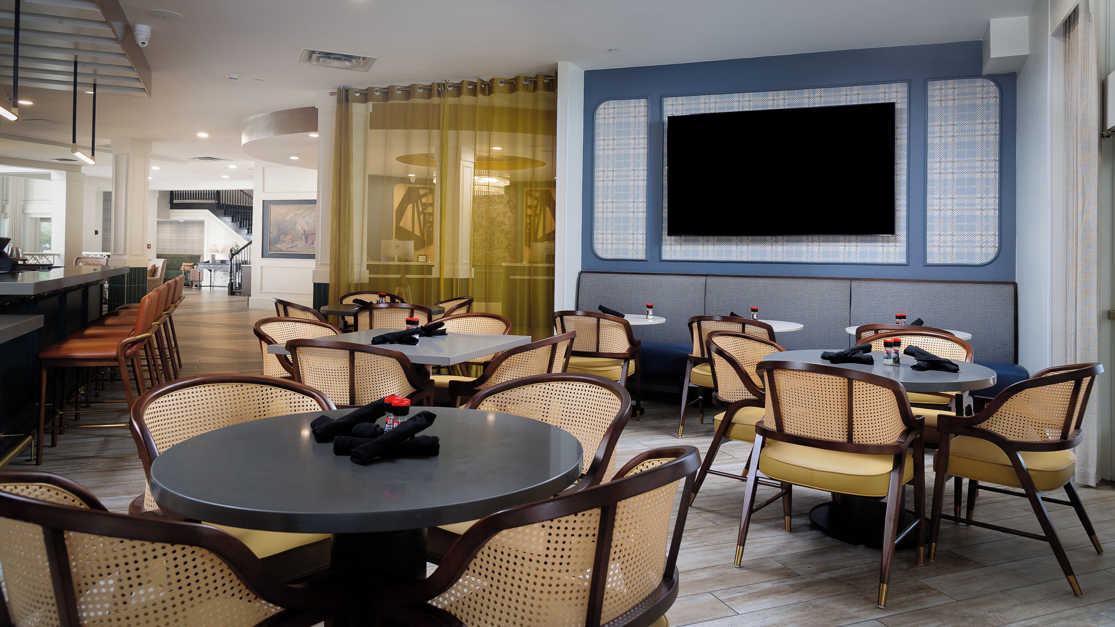 A restaurant with round tables and wicker chairs, a bar area, and a large flat-screen TV mounted on the wall.