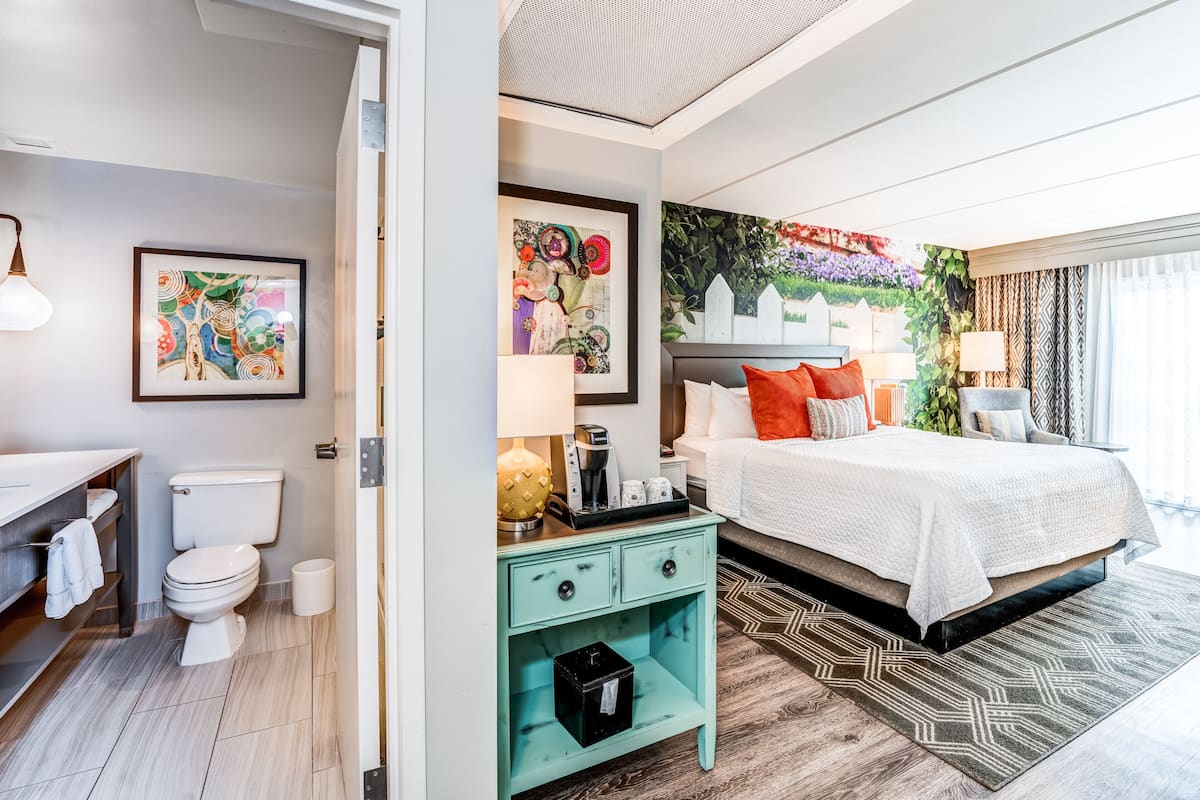 Una habitación de hotel moderna con una cama blanca, una mesita de noche verde, un inodoro blanco y un baño con un lavamanos blanco y una ducha.