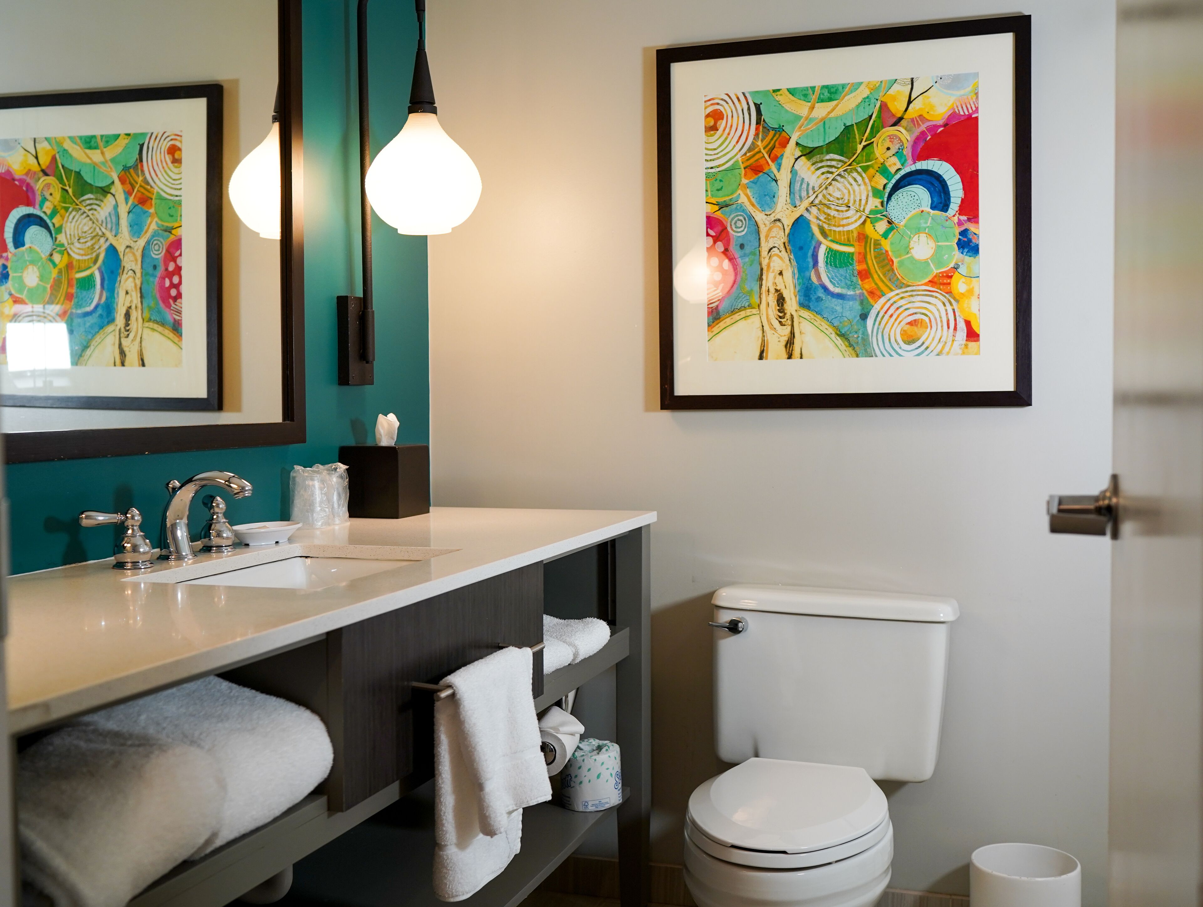 A modern bathroom with a white sink, two hanging light bulbs, a toilet, and two framed abstract paintings on the walls.
