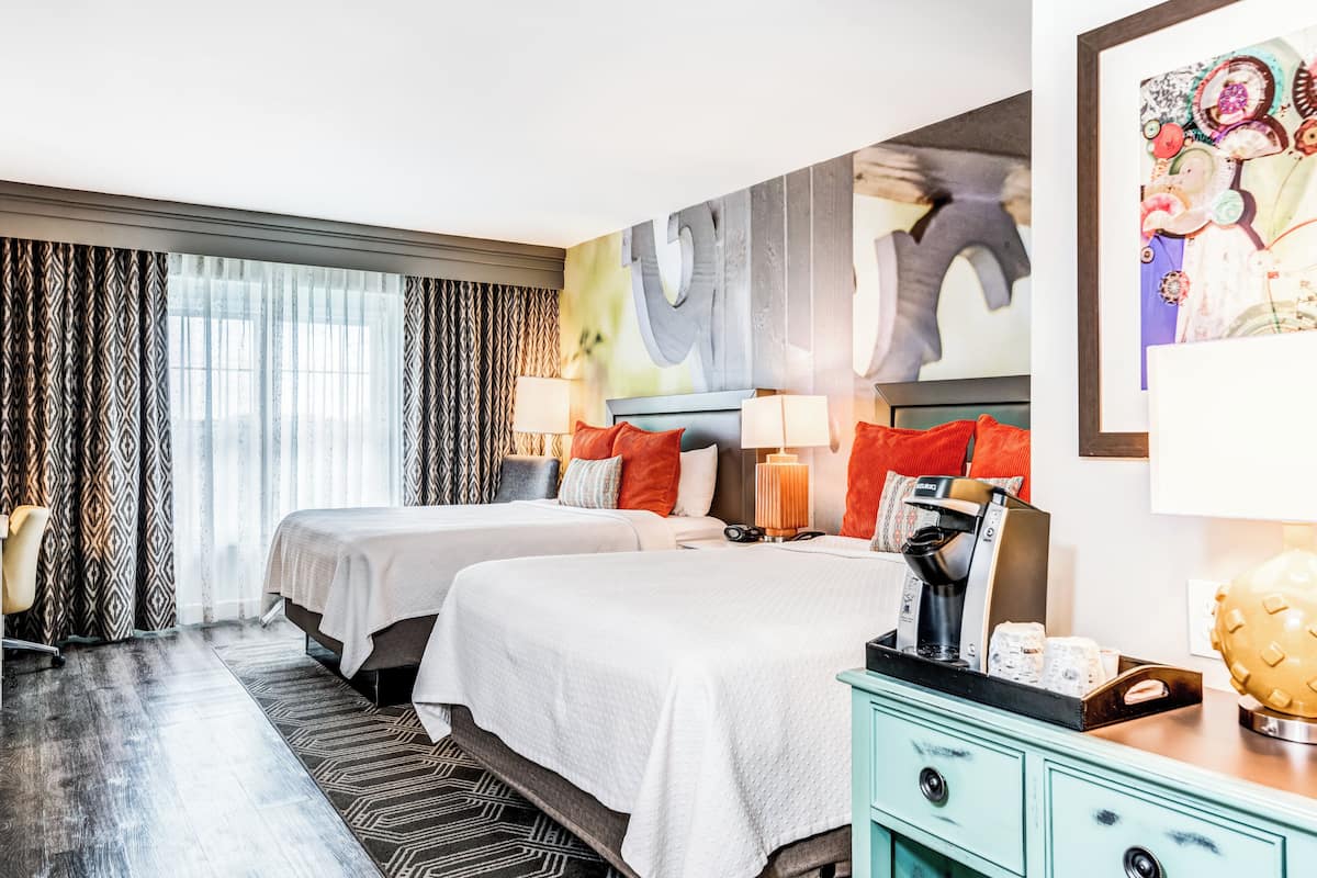 Habitación de hotel con dos camas, un ventanal con cortinas, un escritorio con lámpara y una pared decorada con un cuadro abstracto grande.
