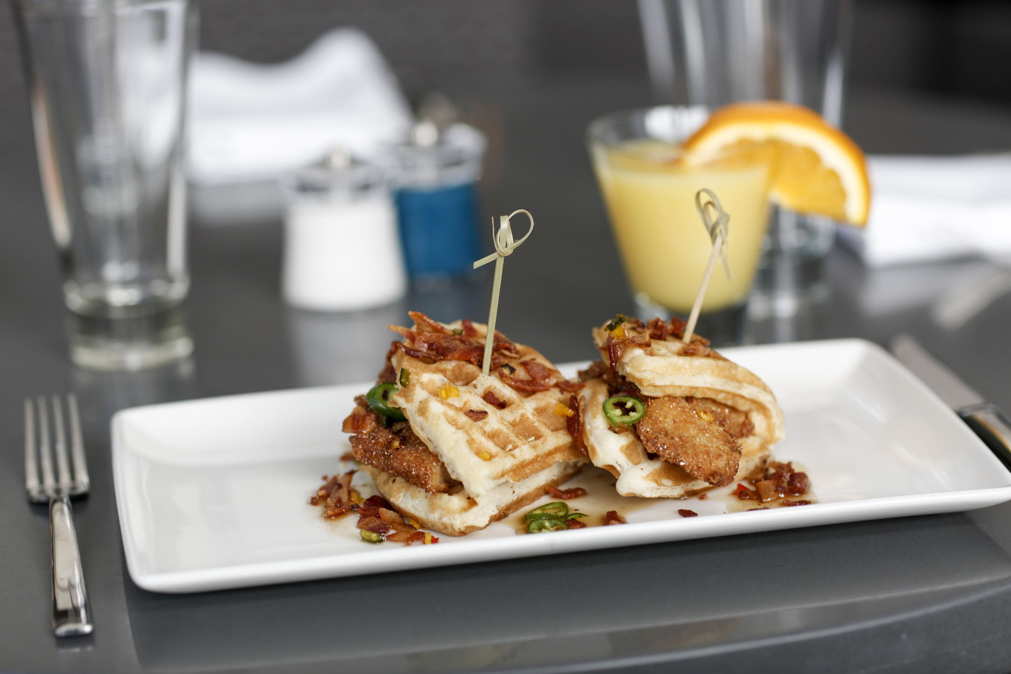 Une assiette de nourriture avec un sandwich coupé en deux, une boisson garnie d’une tranche d’orange et un salier et un poivrier posés sur une table.