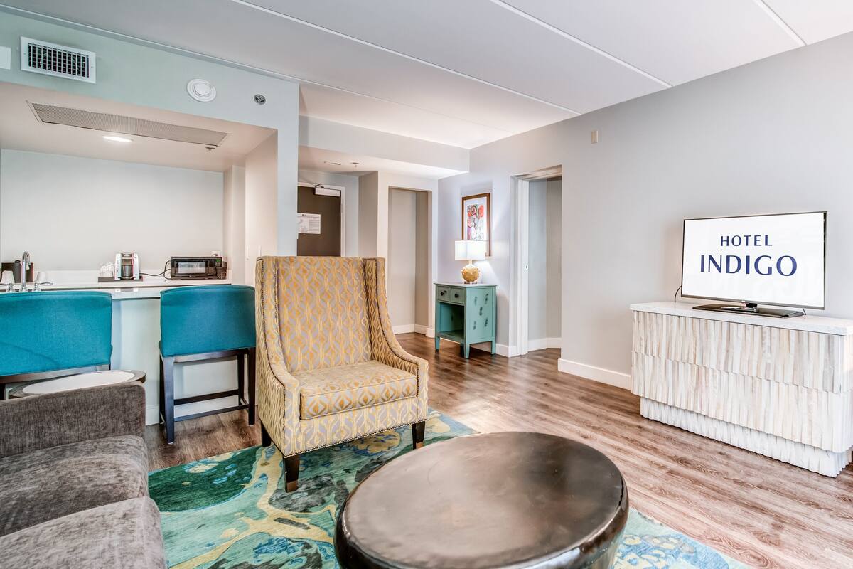 Una habitación de hotel moderna con kitchenette, un sillón estampado, una mesa de centro redonda y un televisor sobre un mueble blanco.