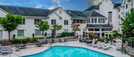 A pool area with lounge chairs and umbrellas, surrounded by a white building with multiple windows and a stone chimney.