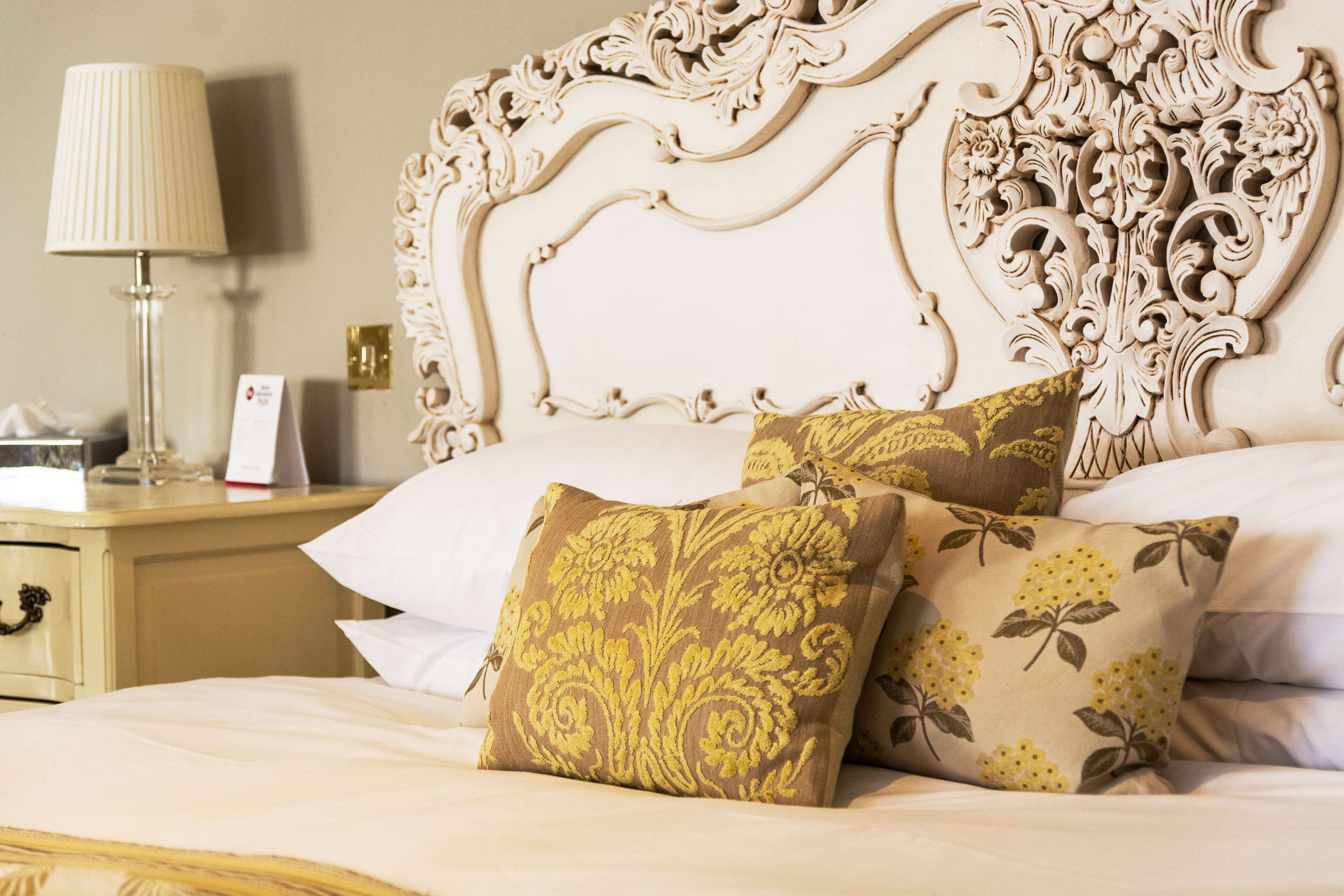 Un lit avec une tête de lit décorative, deux coussins d’ornement, une lampe de chevet et une table de chevet sur laquelle se trouvent une boîte de mouchoirs et un téléphone.