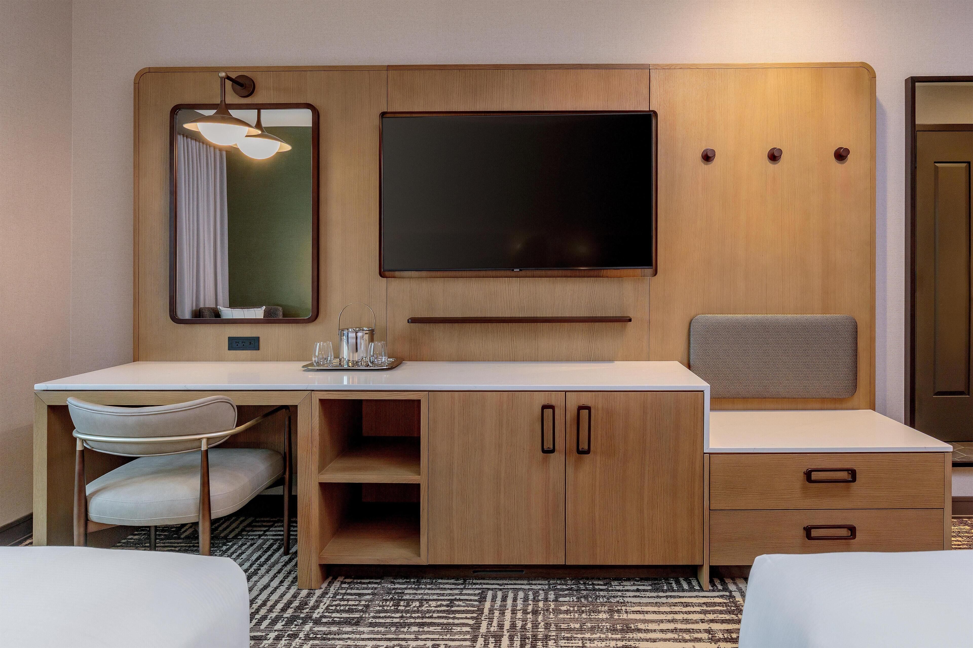 A hotel room with a wooden desk, a flat-screen TV, a mirror, a chair, and a bed.