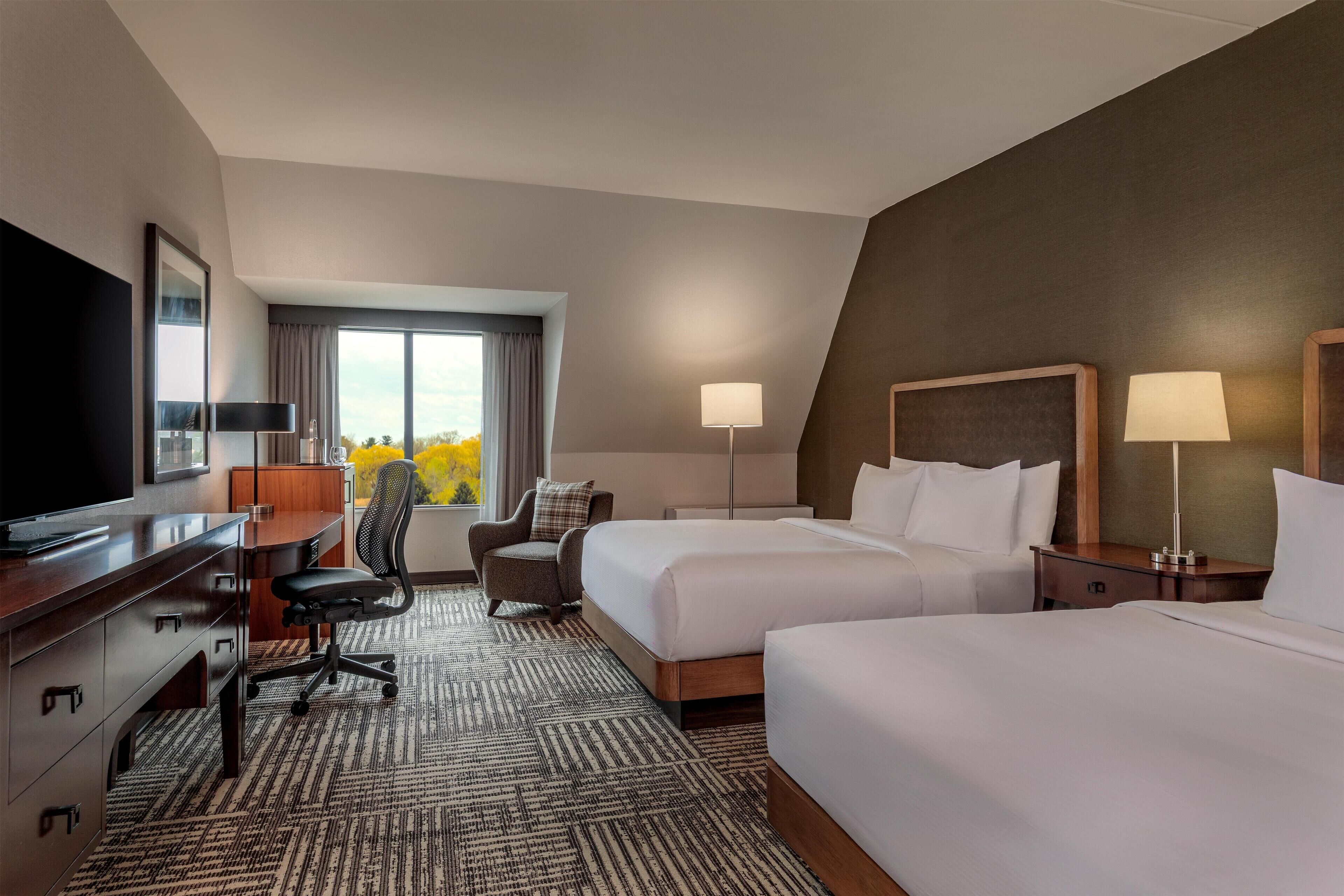 A hotel room with a desk, two beds, a chair, a TV, and a window with curtains.