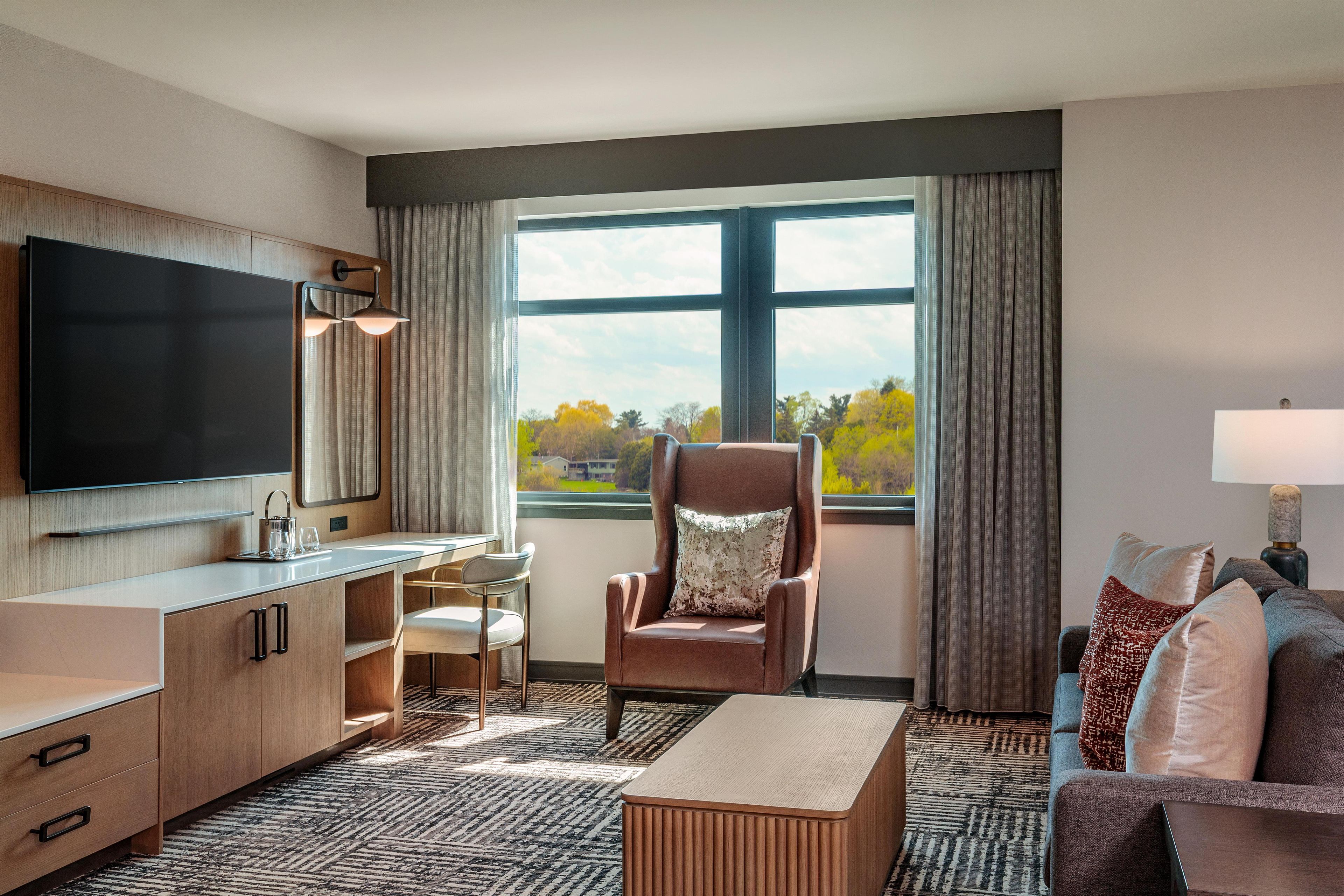 A hotel room with a flat-screen TV, a sofa, a chair, a desk, and a large window with a view.