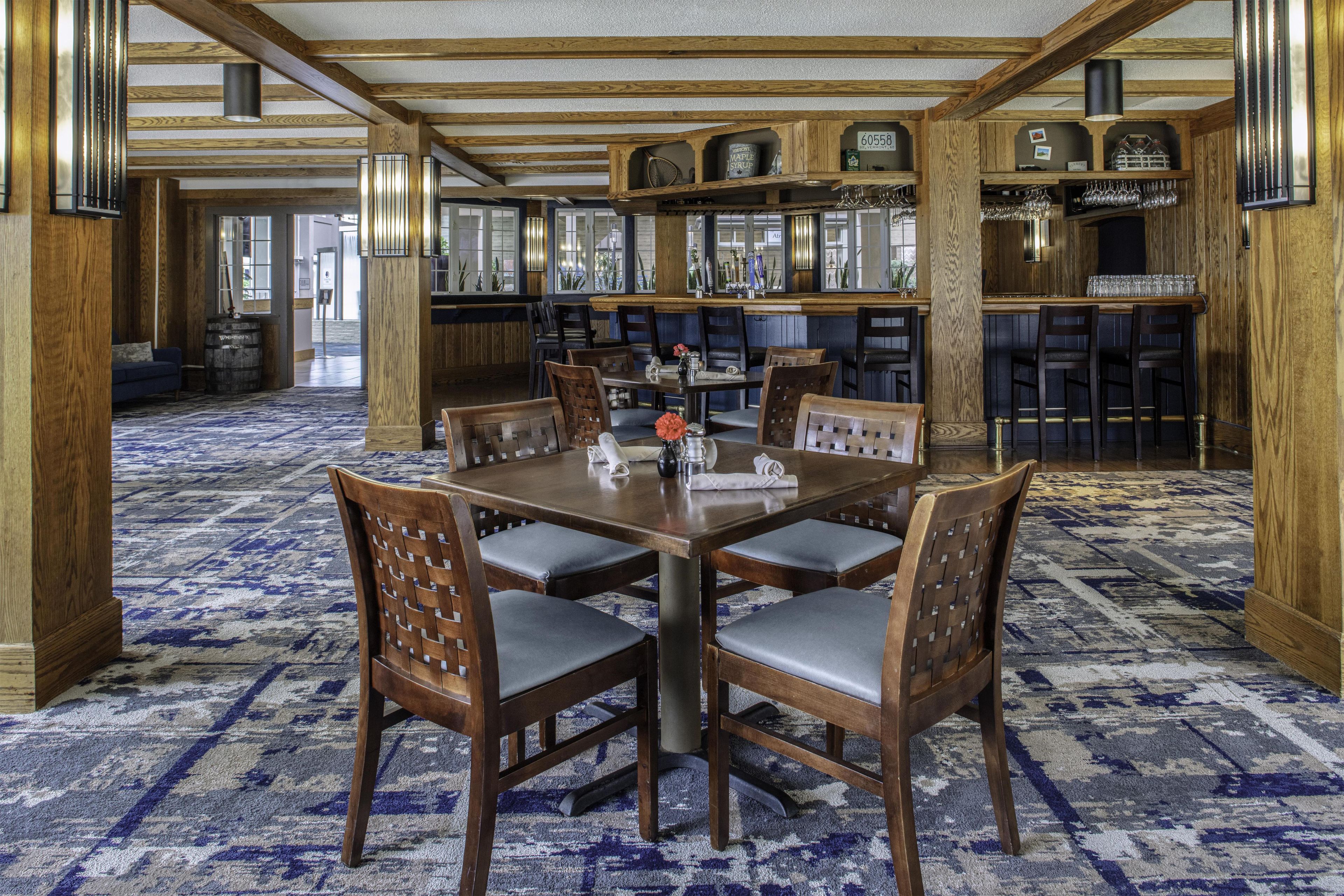 A dining area with a large wooden table surrounded by chairs, a patterned carpet, and a bar area with shelves and glassware.